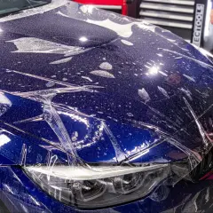 Profesjonalne oklejanie auta folią ochronną na masce BMW. Widać krople wody i pęcherzyki powietrza pod folią.