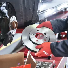 Mechanik w czerwonych rękawiczkach trzyma nową tarczę hamulcową obok zamontowanej tarczy.