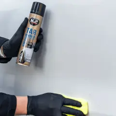 Ręce w czarnych rękawiczkach używają preparatu K2 Tar Remover, by pokazać, jak usunąć naklejki z samochodu. Żółta ściereczka poleruje lakier.