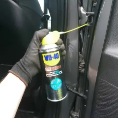 Ręka w rękawiczce aplikuje biały smar litowy WD-40 na zawias drzwi samochodu.