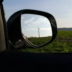 Widok w lusterku samochodowym na wiatrak na tle zielonego pola i nieba.