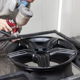 Profesjonalne malowanie felg aluminiowych. Pracownik w masce ochronnej używa pistoletu lakierniczego do aplikacji czarnej farby na felgę.