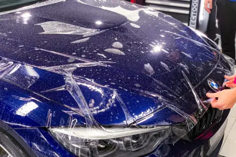 Profesjonalne oklejanie auta folią ochronną na masce BMW. Widać krople wody i pęcherzyki powietrza pod folią.