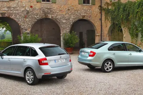 Dwa samochody: srebrny liftback i błękitny hatchback, stoją przed kamiennym budynkiem z łukami i zielenią.
