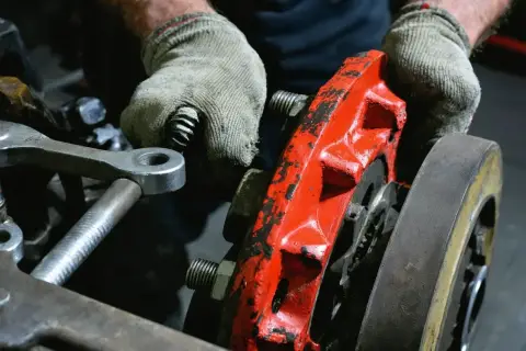 Ręce w rękawiczkach pracują nad czerwonym zaciskiem hamulcowym, przygotowując go do odpowietrzania. To część poradnika "101 napraw odpowietrzanie hamulców".