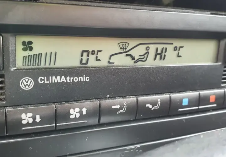 Panel klimatyzacji w samochodzie. Wyświetla 0°C, ikonę nawiewu na szybę i pasażerów, a także ustawienie "HI".