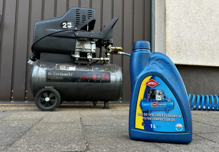 Niebieska butelka z etykietą "OLEJ DO SPRĘŻAREK TŁOKOWYCH PISTON COMPRESSOR OIL" obok sprężarki Einhell.