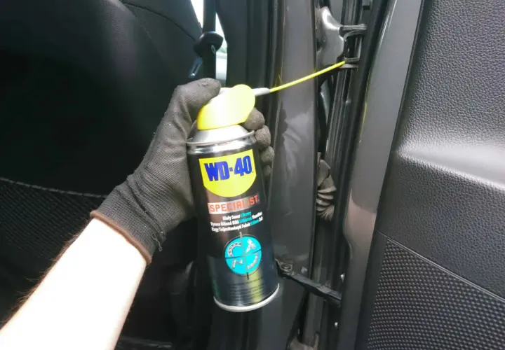 Ręka w rękawiczce aplikuje biały smar litowy WD-40 na zawias drzwi samochodu.