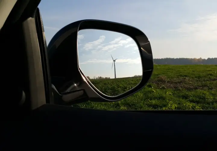 Widok w lusterku samochodowym na wiatrak na tle zielonego pola i nieba.