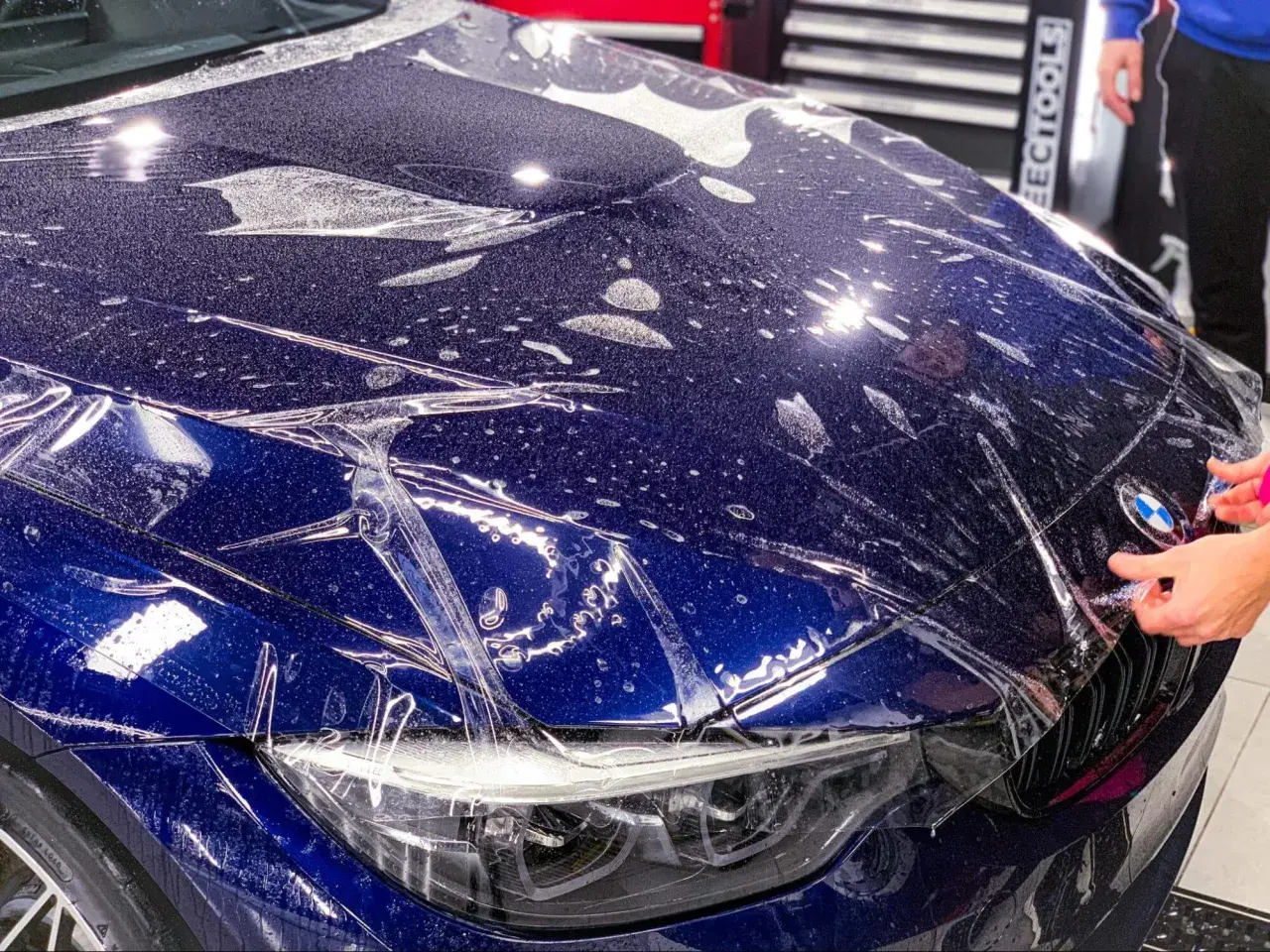 Profesjonalne oklejanie auta folią ochronną na masce BMW. Widać krople wody i pęcherzyki powietrza pod folią.