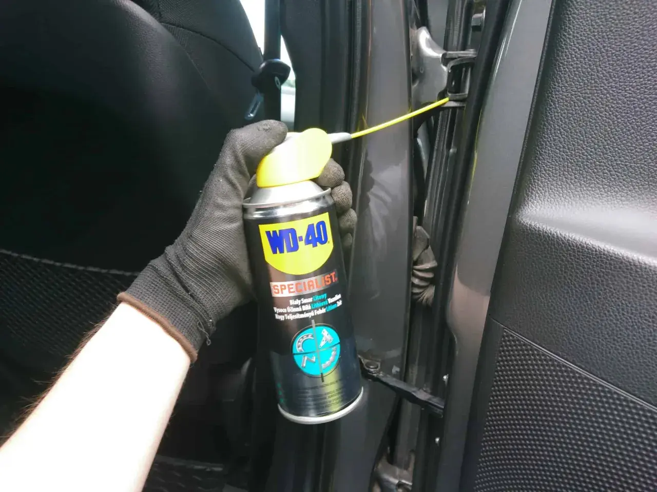 Ręka w rękawiczce aplikuje biały smar litowy WD-40 na zawias drzwi samochodu.