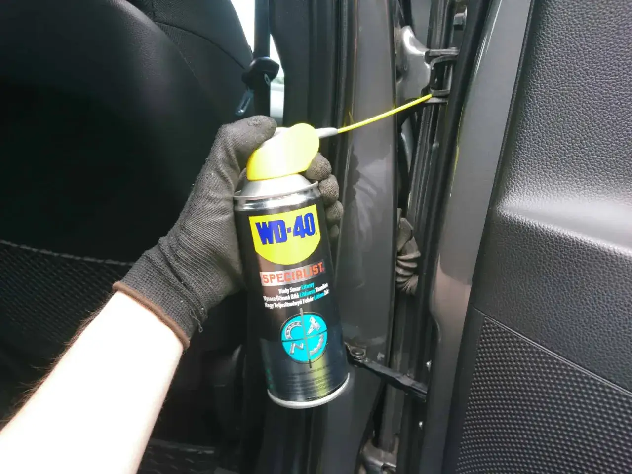 Ręka w rękawiczce aplikuje biały smar litowy WD-40 na zawias drzwi samochodu.