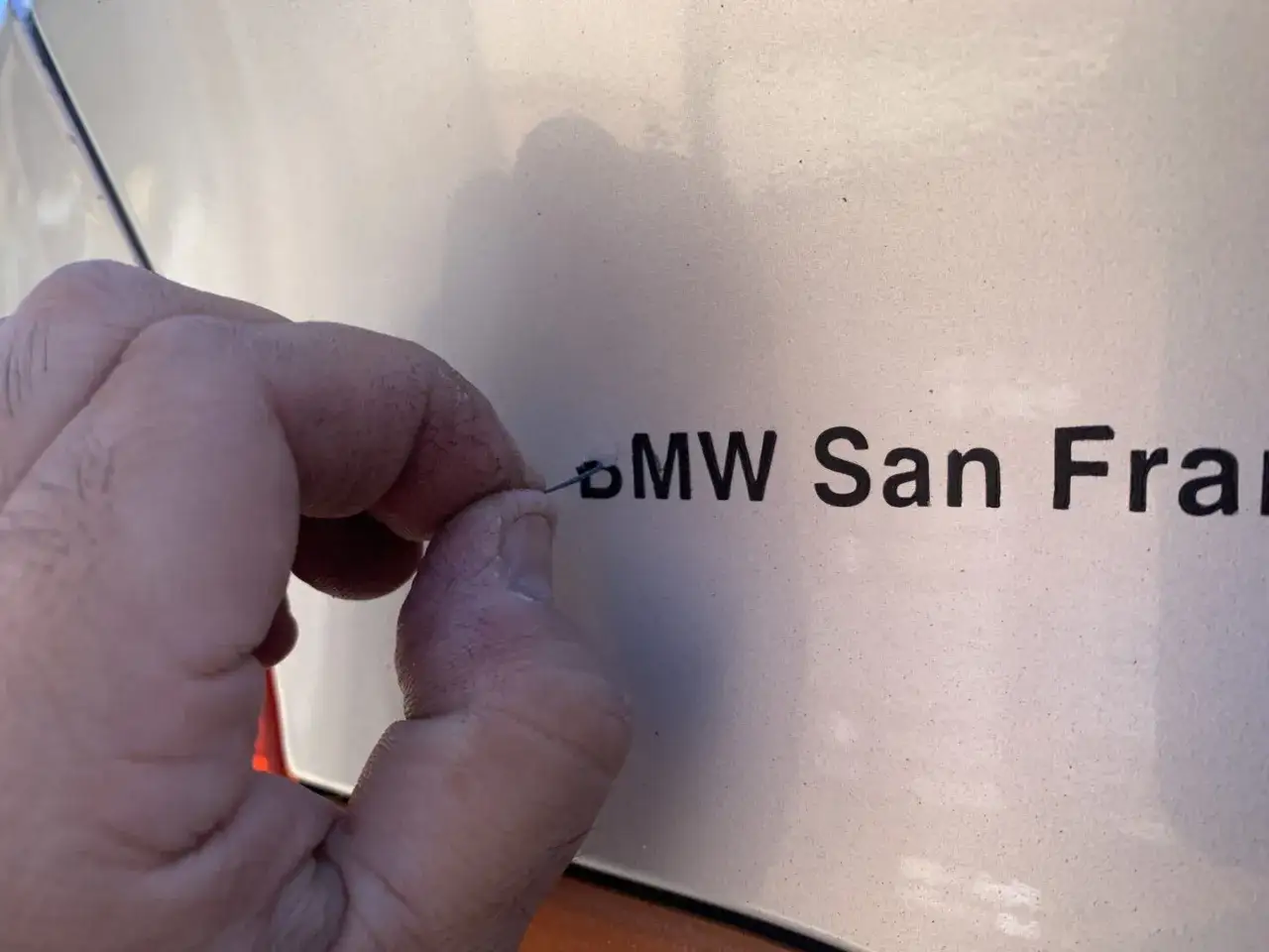 Dłoń z igłą usuwa literę z napisu BMW San Fran. Dowiedz się, jak usunąć naklejki z samochodu.